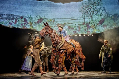 Stage actors perform with two large, realistic horse puppets in an outdoor scene backdrop, with dramatic lighting and expressive poses. A young woman is riding on top of one of the horse puppets.
