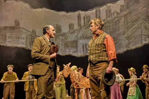 Two men in period costumes talk on stage, holding hats, with a group of performers and a horse puppet behind them in front of a painted city backdrop.