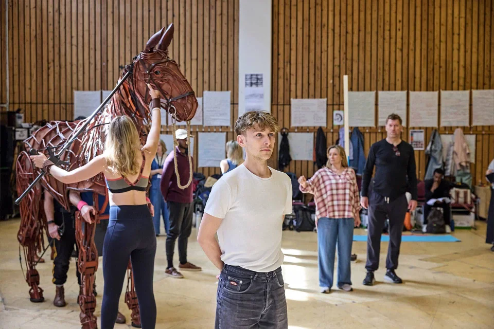 Actors rehearse in a studio; one with a large horse puppet, while others stand or gesture around the room, with notes and coats on the wall in the background.