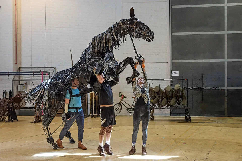 Three people operate a large, rearing horse puppet made of metal and fabric inside a spacious, industrial indoor setting. Military helmets and uniforms are displayed in the background.