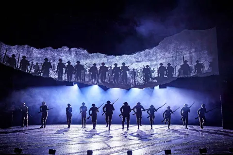 A group of performers with rifles silhouetted on stage, dramatic lighting and a backdrop showing silhouettes of soldiers, enacting a wartime scene.