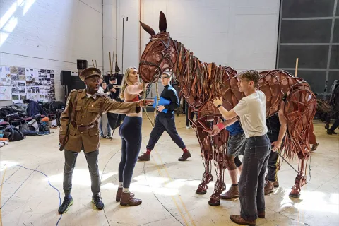 Several people rehearse with a large, lifelike horse puppet in a studio; one person is dressed as a soldier and others help operate the puppet.