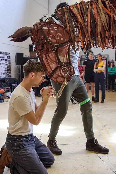 A man kneels and interacts tenderly with a large, realistic horse puppet operated by another person, whilst several people watch in the background.