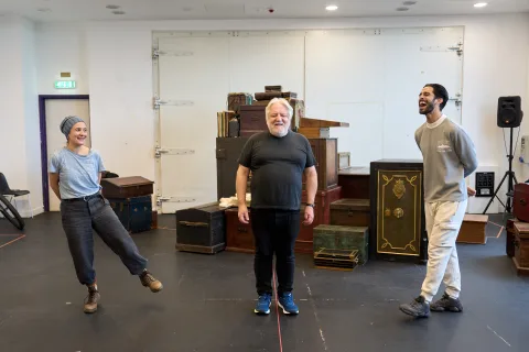 Lyndsey Marshal, Simon Russell Beale and Eben Figueiredo stand in front of a stack of trunks and cases, they laugh together.