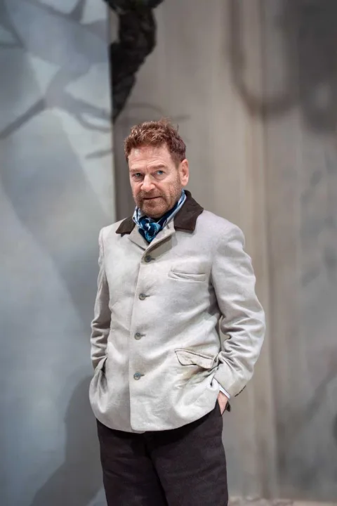 A man wearing a smart light grey jacket with a brown collar stands in the centre of the image with his hands in his trouser pockets. He looks angry.