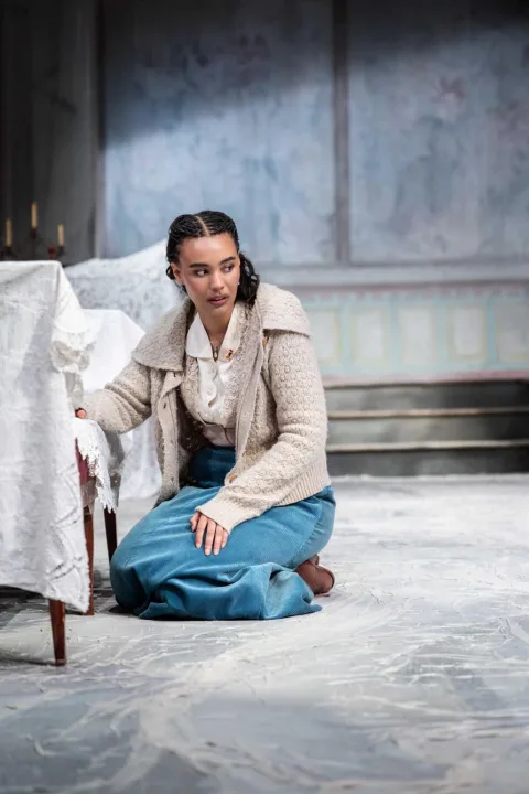 A young woman kneels on a white marbled floor. She rests her arm on a chaise longue covered in a white blanket, she looks sad.