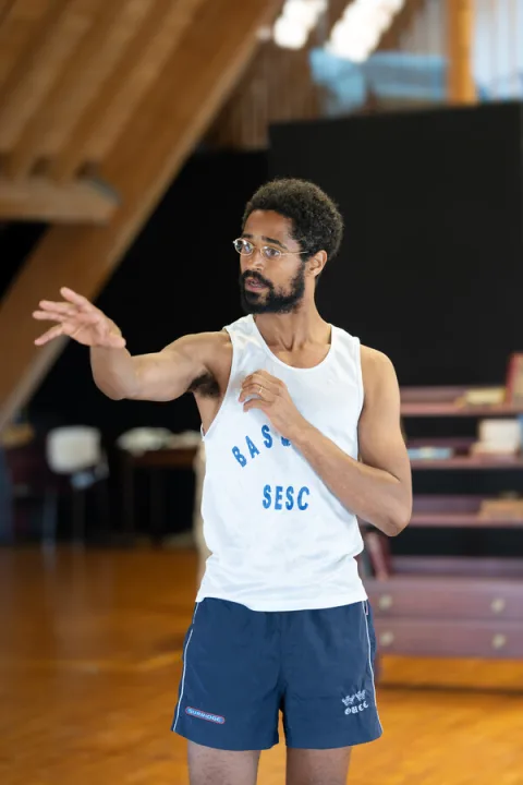 A young man wearing a white vest top and dark blue shorts stands in the centre of the image with his hands stretched forwards.