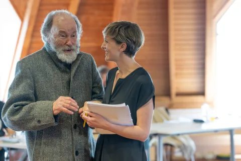 An older man wearing a buttoned up grey jacket stands next to the Director having a conversation.