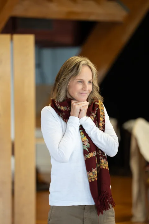 A woman wearing a white long-sleeved top and red scarf stands in the centre of the image smiling with her hands clasped in front of her.