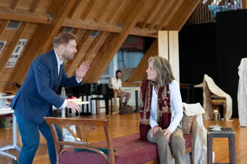 A man wearing a dark blue suit stands opposite a woman wearing a long sleeved white top sitting on a chair.