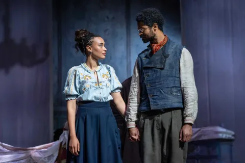A young man and woman stand next to each other holding hands. The woman wears a light blue shirt with yellow flowers on and the man wears a dark blue waistcoat and grey trousers. They look into each other’s eyes.