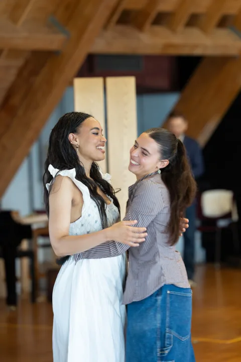 A young woman wearing a white dress stands opposite another young woman in a grey stripy shirt. They have their arms round each other in a hug.