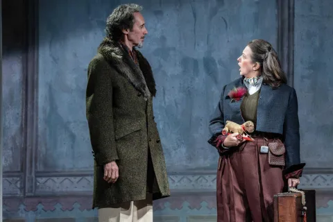 A man wearing a grey coat with a fur collar and a pair of grey trousers stands in the centre of the image. A woman stands opposite him wearing a short blue jacket and red shiny trousers. She carries a small toy dog and looks shocked.