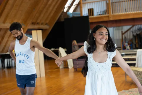 A young man wearing a white vest top and a young woman wearing a white dress stand in the centre of the image holding hands.