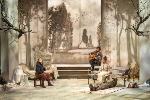 A woman sits on a stool o the left of the image. A man playing a guitar sits opposite her on a bench next to a woman. A man lies on a picnic blanket on the floor. The Cherry Orchard is behind them.