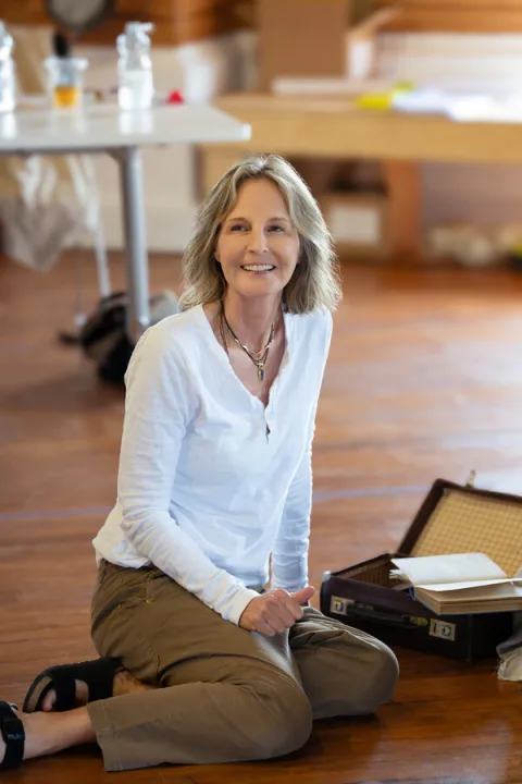 A woman wearing a white long sleeve top and grey trouser sits on the floor next to an open suitcase.