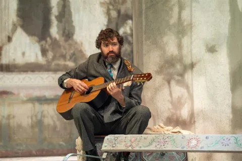 A young man sits in the centre of the image playing an acoustic guitar. He wears a grey suit and looks sad.