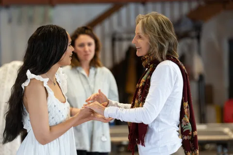 A young woman wearing a white dress stands opposite an older woman wearing a long-sleeved white shirt and a red scarf.