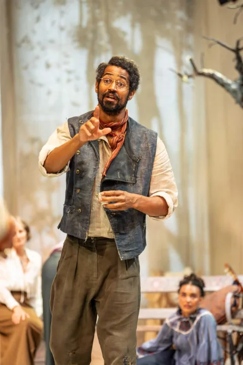 A young man wearing a blue waistcoat and grey trousers stands in the centre of the stage. He wears glasses and is talking to someone out of shot. His left arm is stretched outwards.