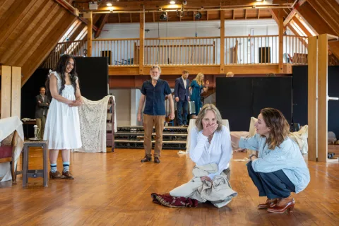 A group of people stand in a wooden rehearsal room running through lines.