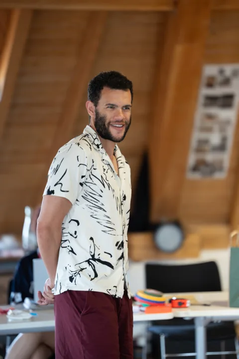 A young man wearing a patterned black and white shirt and dark red trousers stand sin the centre of the image smiling.