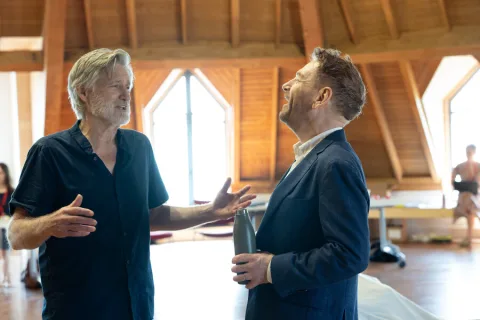 A man wearing a dark blue suit and a man wearing a dark blue shirt stand opposite each other laughing and having a conversation.