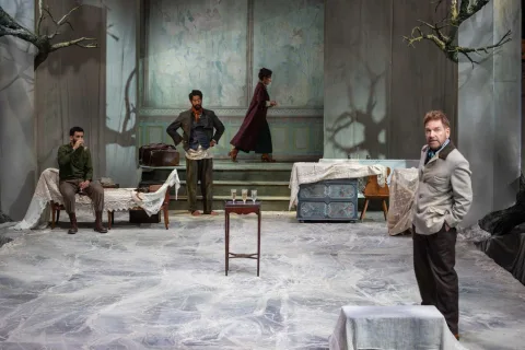 A group of four people stand in a room with furniture covered in blankets. A tray of champagne flutes is on a table I the centre and one man drinks a glass of champagne.