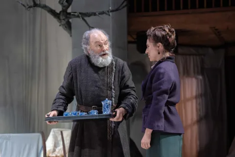 An old man wearing a floor length brown tunic carries a tray with a white and blue pattered China tea set on. A young woman wearing a blue jacket and grey trousers stands next to him.