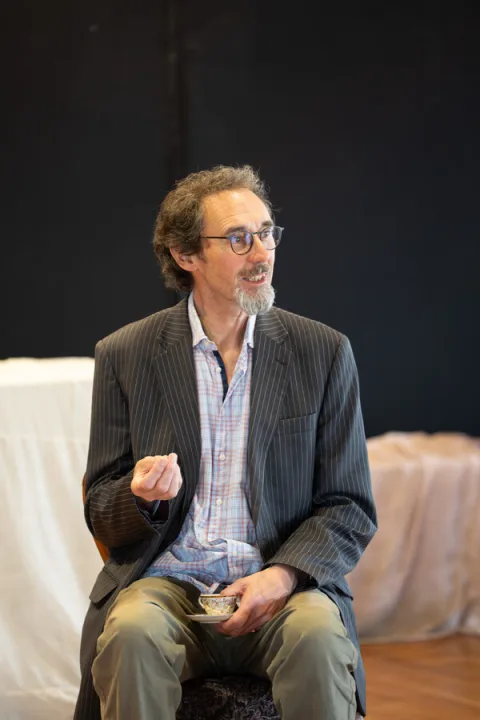 A man wearing dark green trousers and a striped jacket sits on a chair holding a small teacup and saucer.
