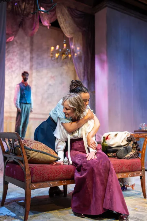 A woman wearing a white shirt and a light purple shirt sits on a chaise longue in the centre of the image. A young woman stands behind her and wraps her arms round her in a hug, smiling. A young man stands in the background.