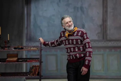 A man with a white beard and wearing a red and white jumper and yellow necktie rests his right hand on a bookcase. He is smiling and laughing.