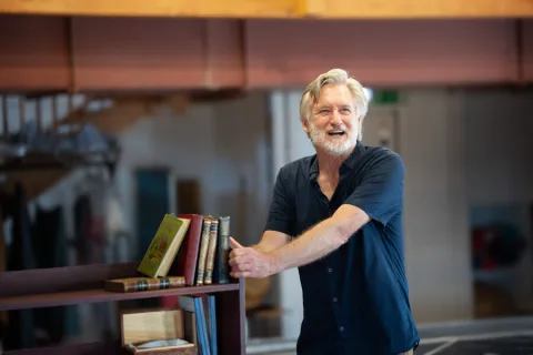 A man wearing a dark blue shirt stands next to a bookcase smiling.
