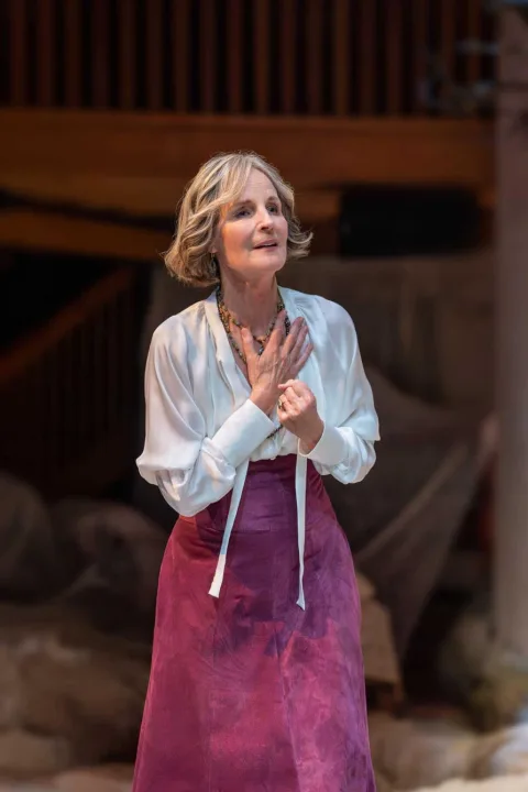 A woman wearing a light purple skirt and a white shirt stands in the centre of the image. She holds her left hand to her chest and speaks to someone out of shot.
