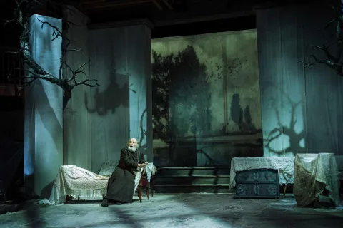 An old man sits on a chaise longue in the centre of a dark room looking frightened. All of the furniture in the room is covered with blankets. I the background is the cherry orchard.