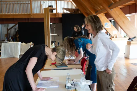 A woman wearing a long-sleeved white top stands opposite the Director who’s reading a script. Both women are laughing.