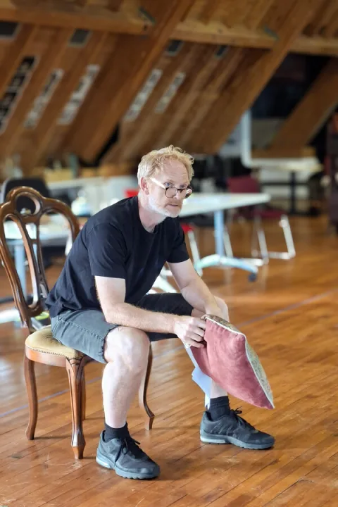 A man wearing a dark blue t-shirt and grey shorts sits on a wooden chair. He looks towards someone out of shot and holds a red cushion.