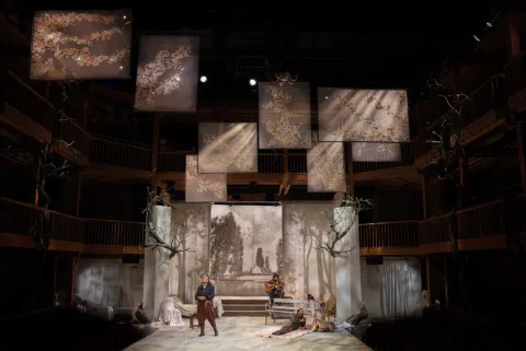 A group of people stand on stage in the centre of the image behind them is a backdrop with the cherry orchard on it. There are eight silk screens suspended above the stage with cherry blossom on.