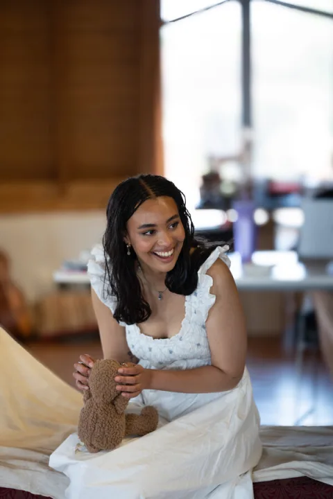 A woman wearing a white dress sits on a chair holding a small brown teddy bear.