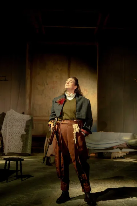 A woman wearing a pair of red shiny trousers, a green jumper and a short blue jacket with a red feather stands in the centre of the image. She holds a rifle in her left hand and has her right hand in her pocket.