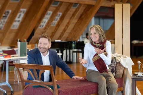 A man wearing a dark blue shirt kneels down behind a red chair, a woman wearing a long-sleeved white top and red scarf sits on the chair.