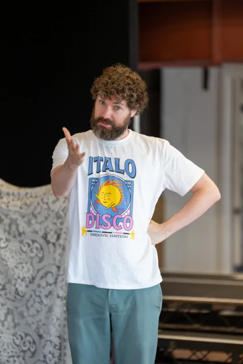A man wearing a white t-shirt and blue trousers stands in the centre of the image with his left arm out-stretched.