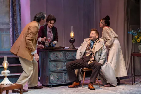 A group of three people stand round a man sat on a wooden chair. A large blue set of drawers is behind them with an oil lamp on the top. A woman stands behind and pours glasses of water from a jug.
