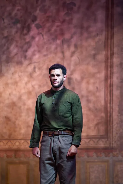 A young man wearing a dark green shirt and grey trousers stands in the centre of the image. He has his fists clenched by his side and looks sad. A wall with brown wallpaper on is behind him.