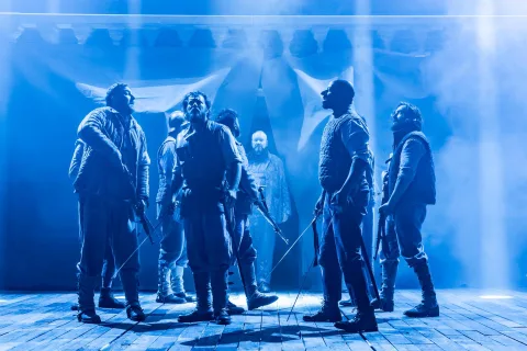 A group of men stand in the centre of the stage illuminated by a blue light. They carry swords.