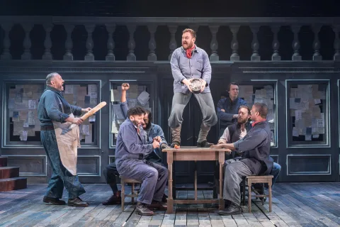 A group of men sit round a table on benches. One of the men stands o the large wooden table and another man stands at the side wearing a grubby apron and carrying a baguette.