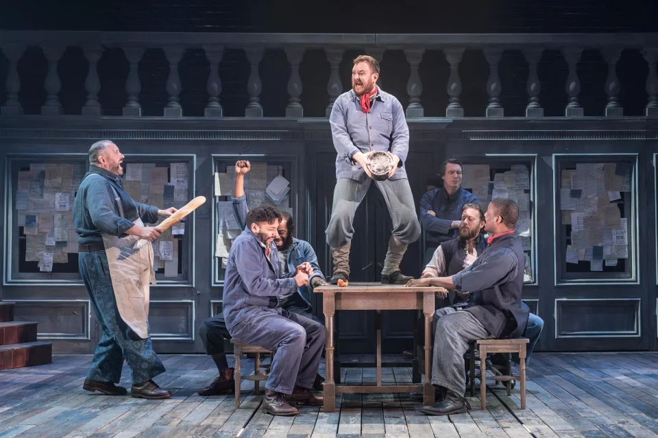 A group of men sit round a table on benches. One of the men stands o the large wooden table and another man stands at the side wearing a grubby apron and carrying a baguette.