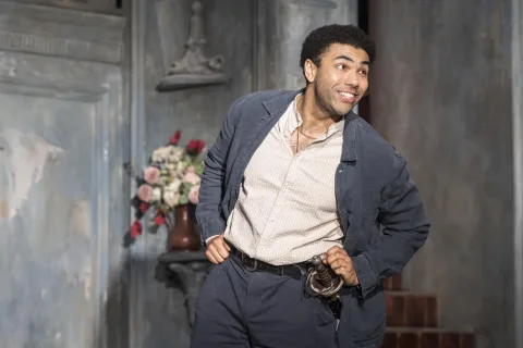 A young black man stands in the centre of the image smiling. He wears a grey jacket, a white linen shirt and a sword hanging from his belt.
