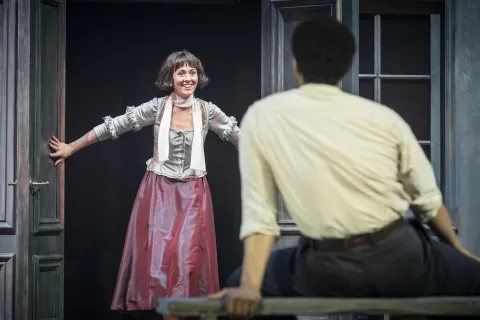 A woman wearing a red silk skirt and grey silk top stands in a doorway. A young man sits opposite her watching her enter.
