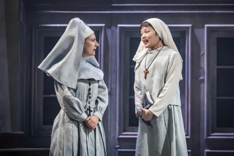 Two young women dressed as nuns stand in the centre of the image talking to each other. One holds a bible.