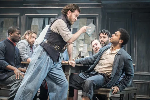 A man wearing a black waistcoat and blue trouser stands in the centre of the image holding a notebook toward another man sat at a table.